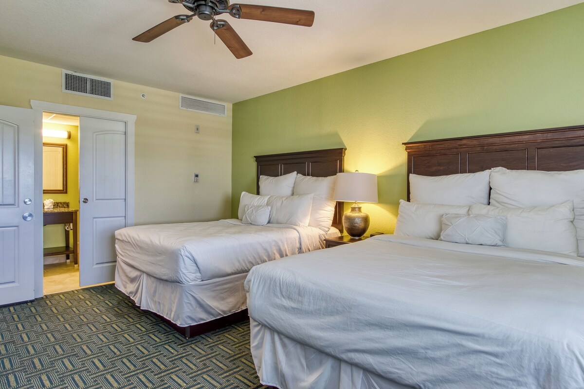 A hotel room with two beds, a ceiling fan, a nightstand with a lamp, and a bathroom visible through an open door.