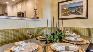 A dining area with a table set for four, featuring woven placemats, white plates, and wine glasses. A framed picture of a beach scene hangs on the wall.