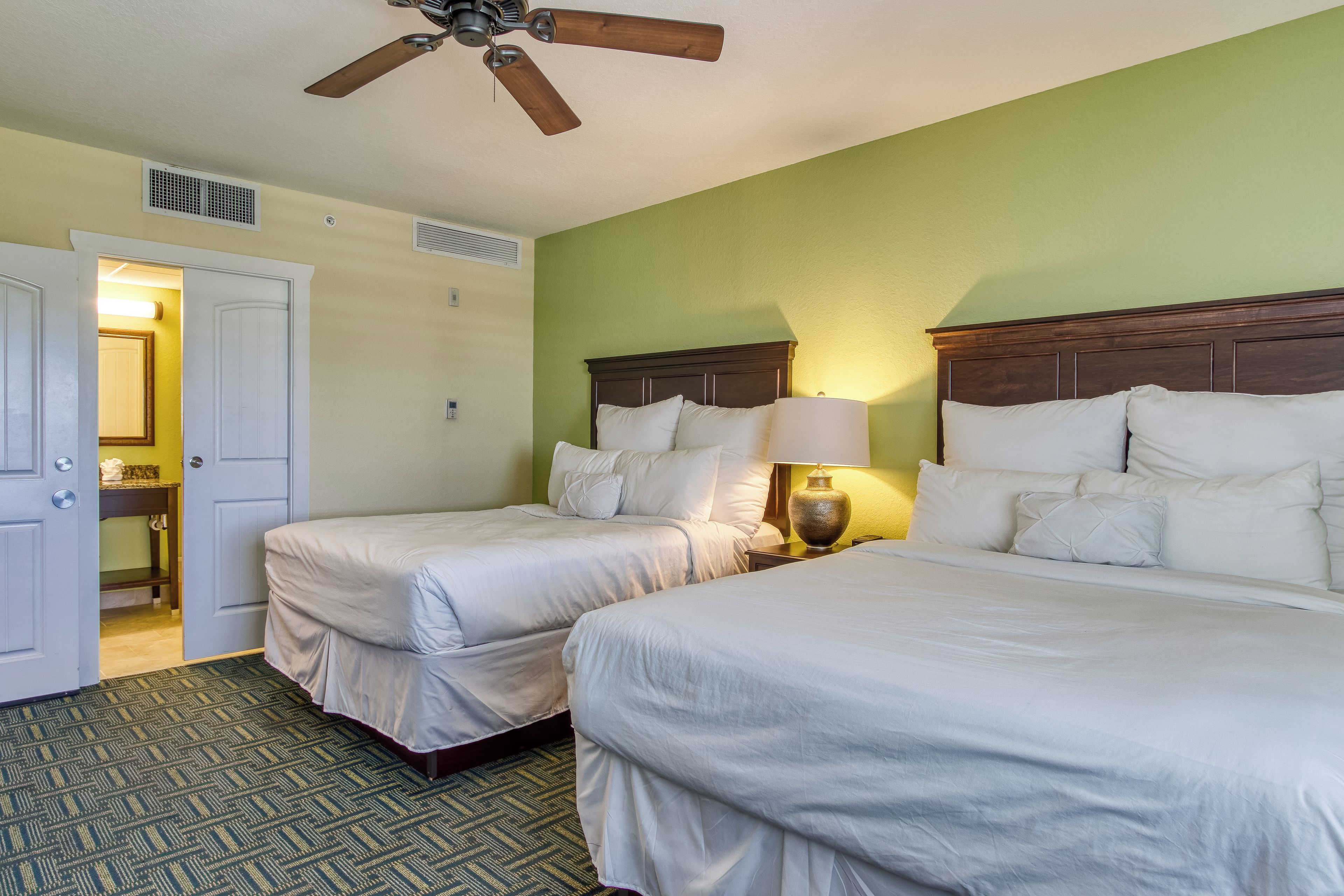 Quarto de hotel com duas camas, um ventilador de teto, mesa de cabeceira com abajur e banheiro visível por uma porta aberta.