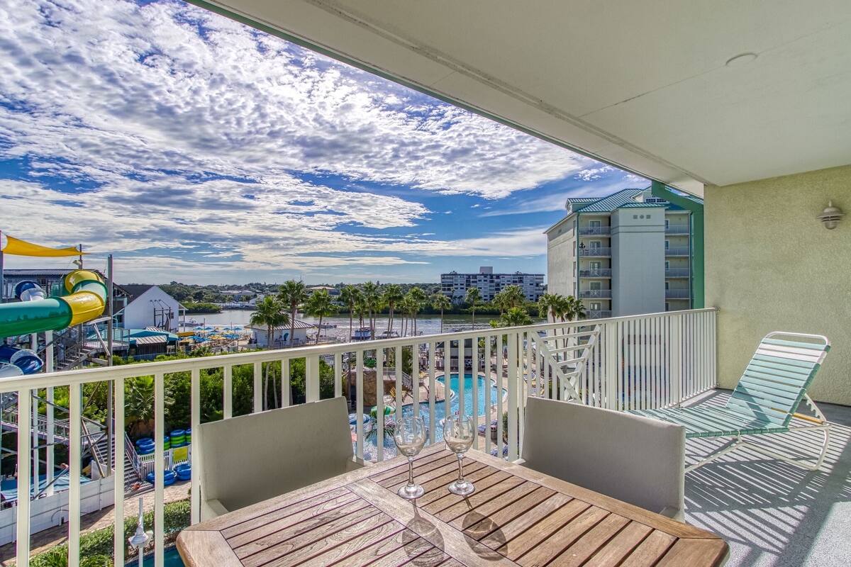 A balcony with a view of a pool area, a table with two glasses, and a chair.
