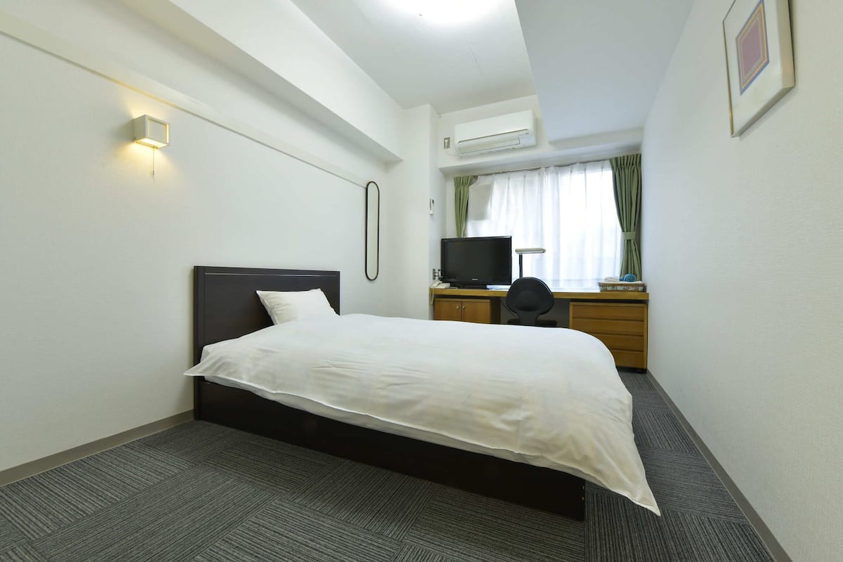 A hotel room with a bed, a desk with a computer, a television, and an air conditioning unit.