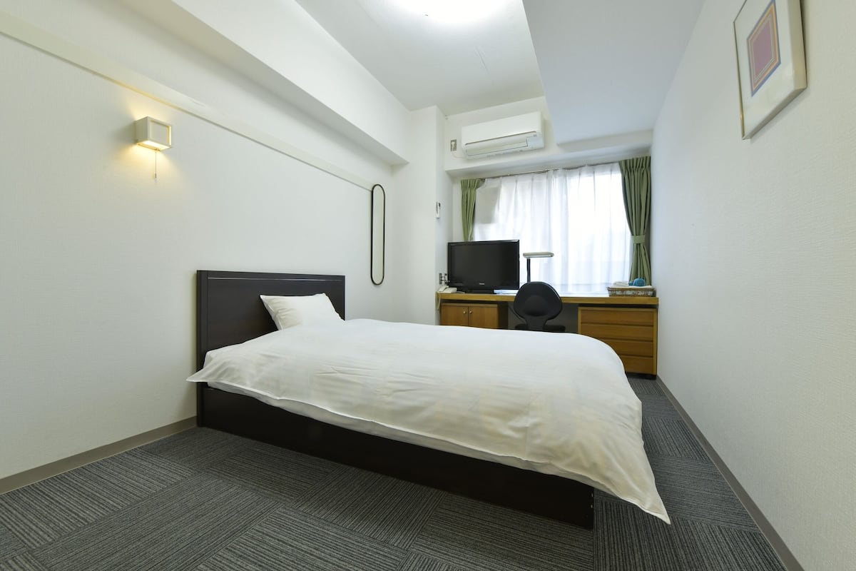 A hotel room with a single bed, a desk with a chair, a television, and an air conditioning unit.