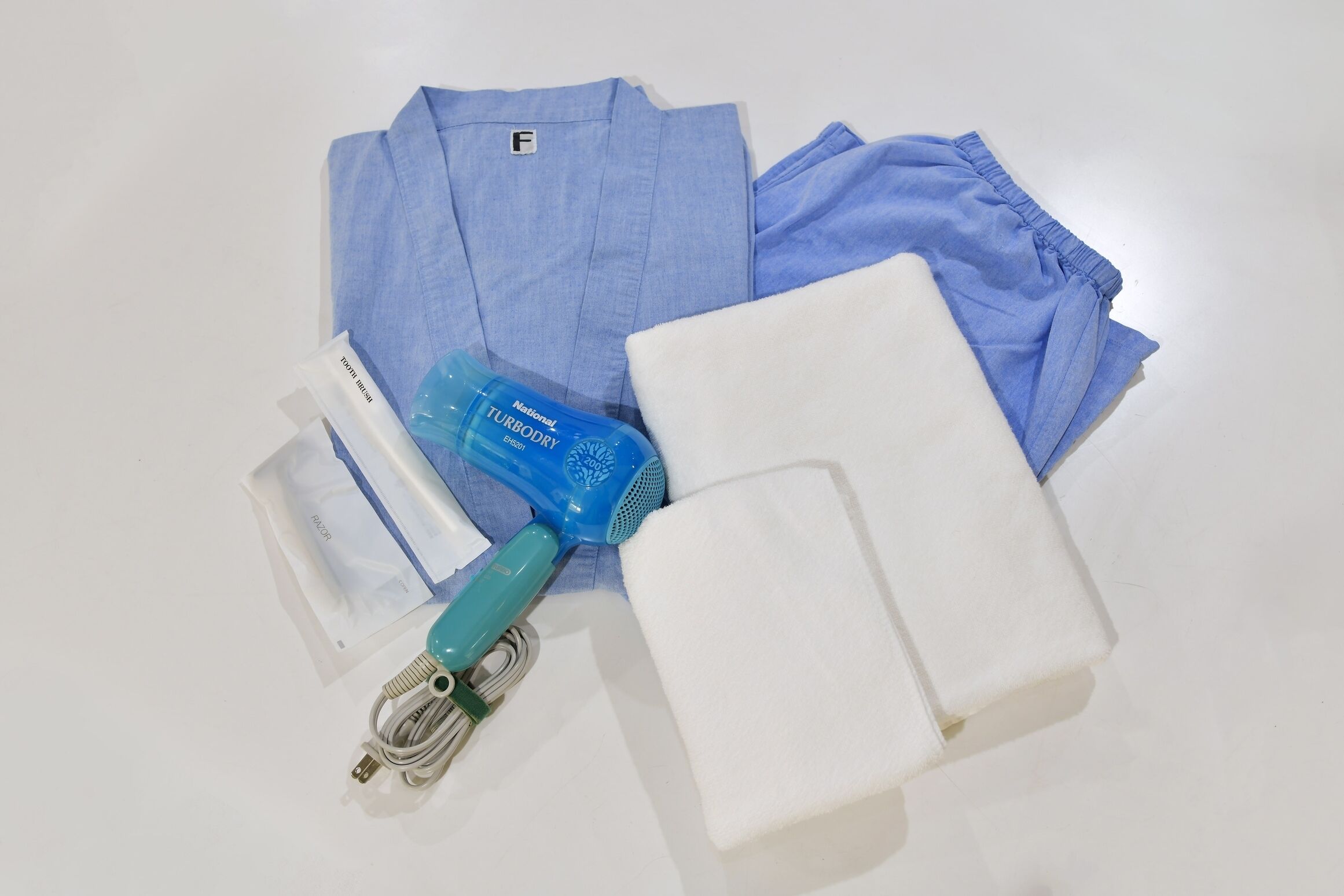 A neatly folded blue shirt, a white towel, a blue hairdryer, and a white charging cable on a white surface.