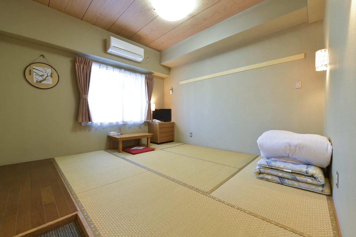 A room with tatami flooring, a futon bed, a small wooden desk, a TV, and a wall-mounted air conditioner.
