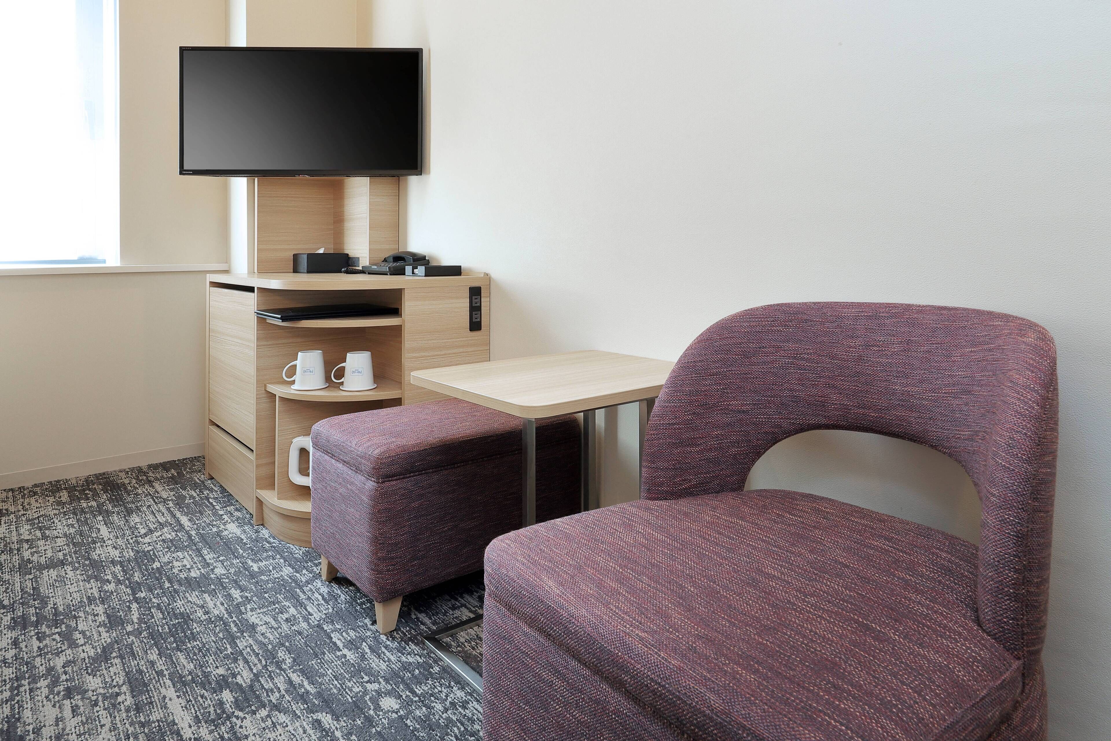 A hotel room with a bed, a desk with a flat-screen TV, a small table, and a purple chair.