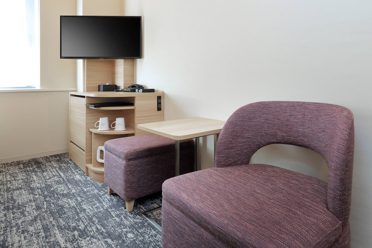 A hotel room with a bed, a desk with a flat-screen TV, a small table, and a purple chair.