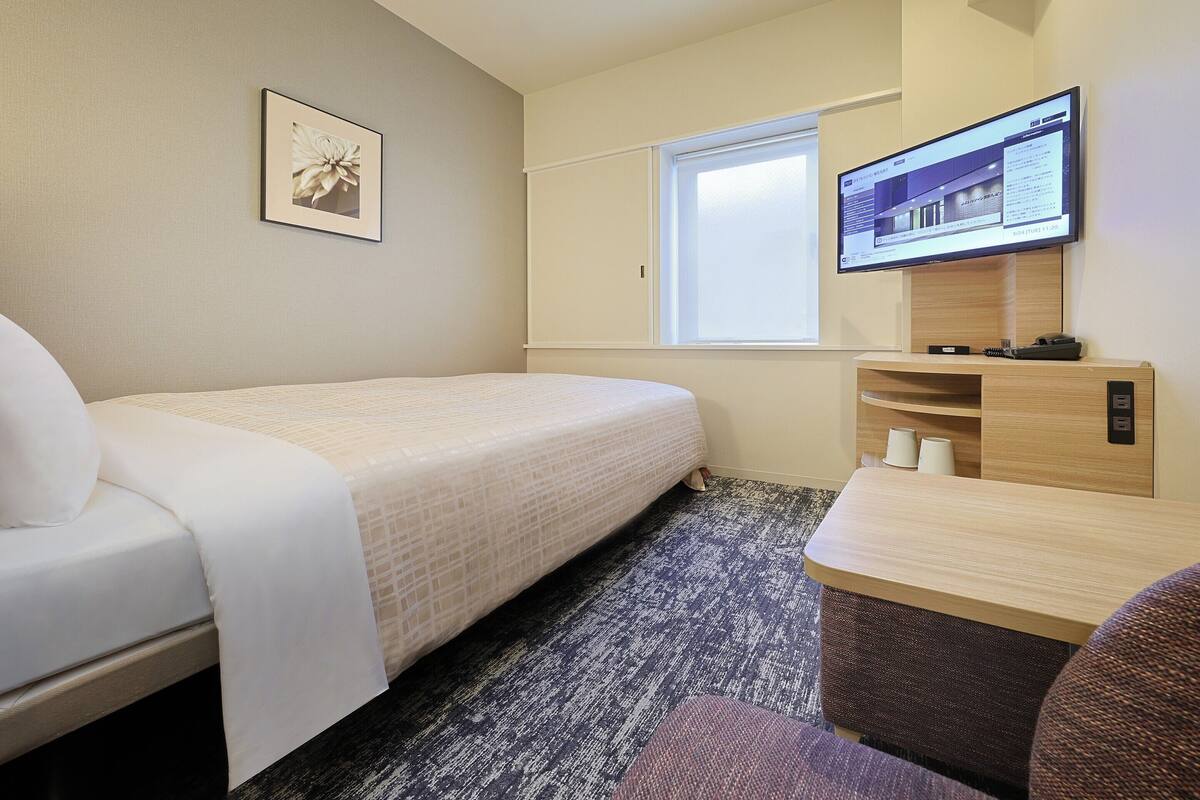 A hotel room with a single bed, a desk with a computer, a television mounted on the wall, and a window.