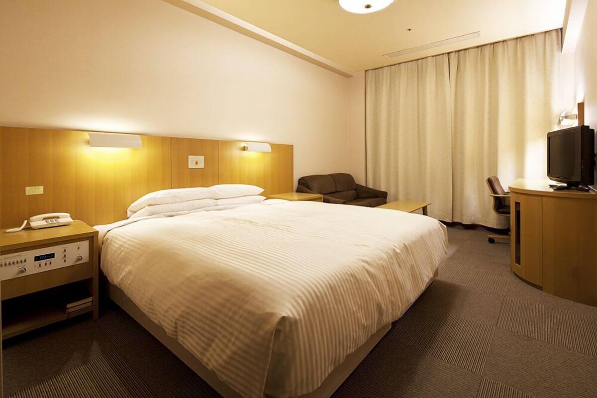 A hotel room with a large bed, bedside tables, a desk with a chair, a television, and curtains.