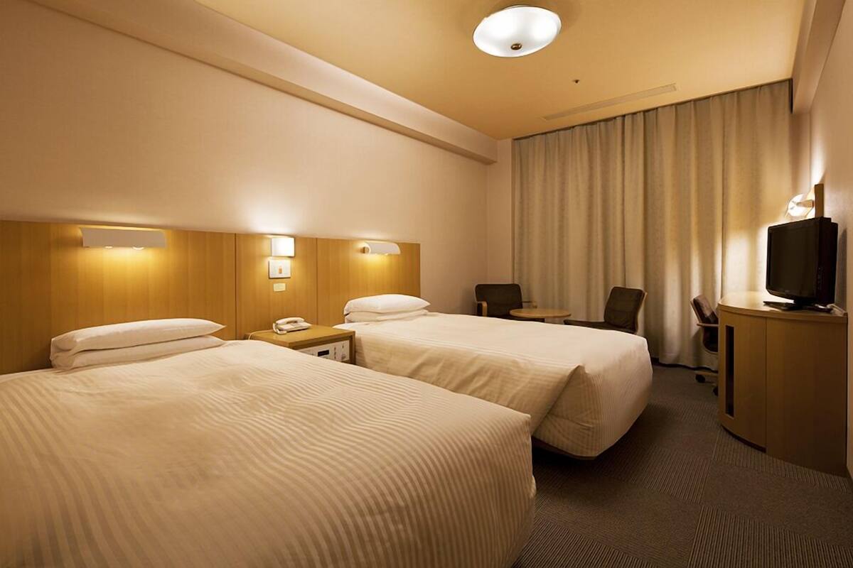 A hotel room with two beds, a desk, a chair, a television, and a window with curtains.
