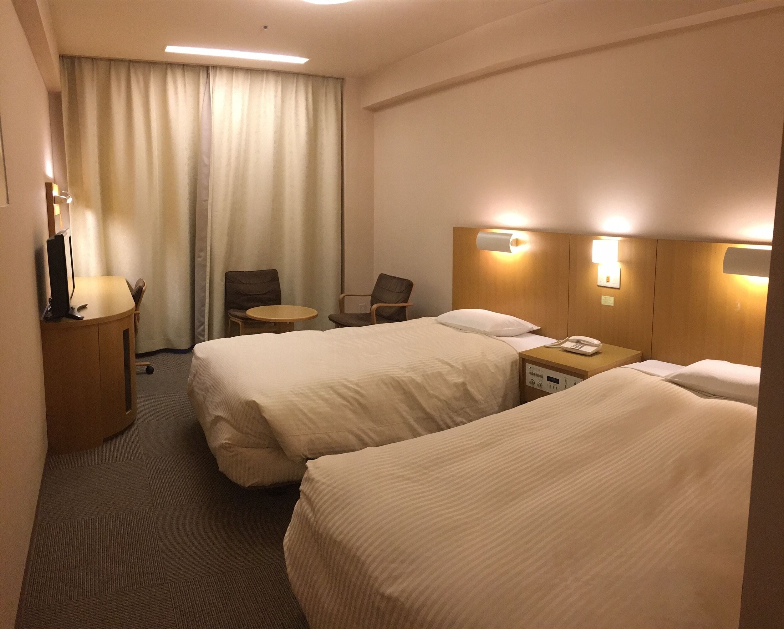 A hotel room with two beds, a desk, a chair, a small table, and a curtain.