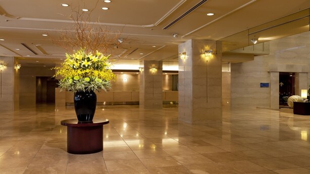 En rymlig entréhall med en stor vas med gula blommor på ett piedestal, en receptionsdisk och en modern takdesign.