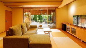 A modern living room with a sofa, a television, and a view of a garden through large sliding doors. - Kaikatei Hanagozen (Kyotango)