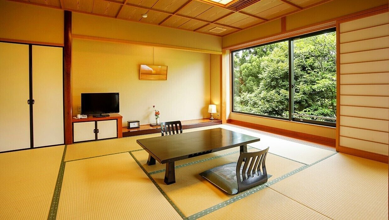 Una habitación con tatamis, una mesa baja, sillas, un televisor y una ventana con vistas a árboles.
