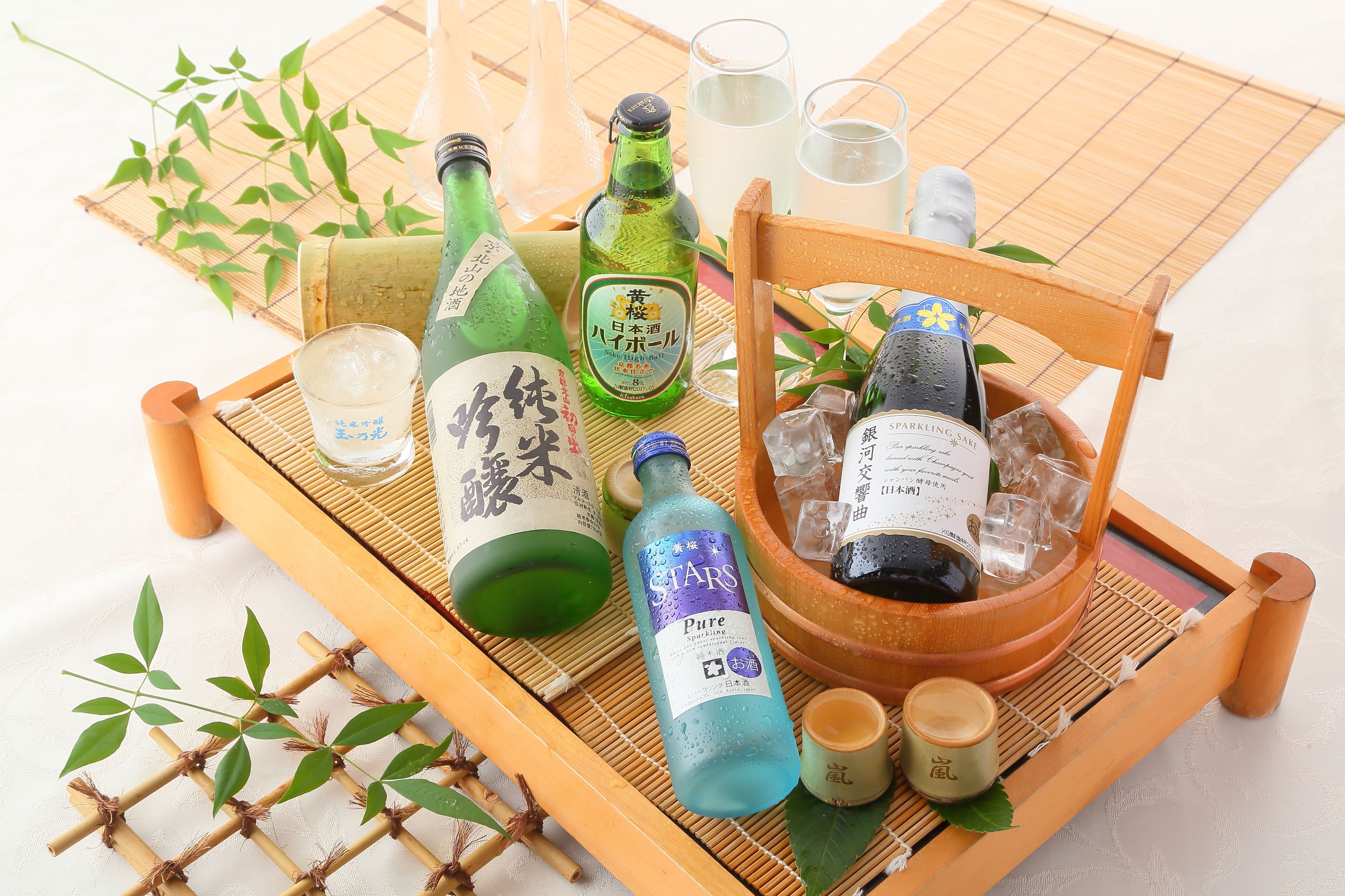A wooden tray with various bottles and glasses, including a green bottle with Japanese characters, a blue bottle labeled "STARS Pure," and a wooden bucket with a bottle of sparkling wine.