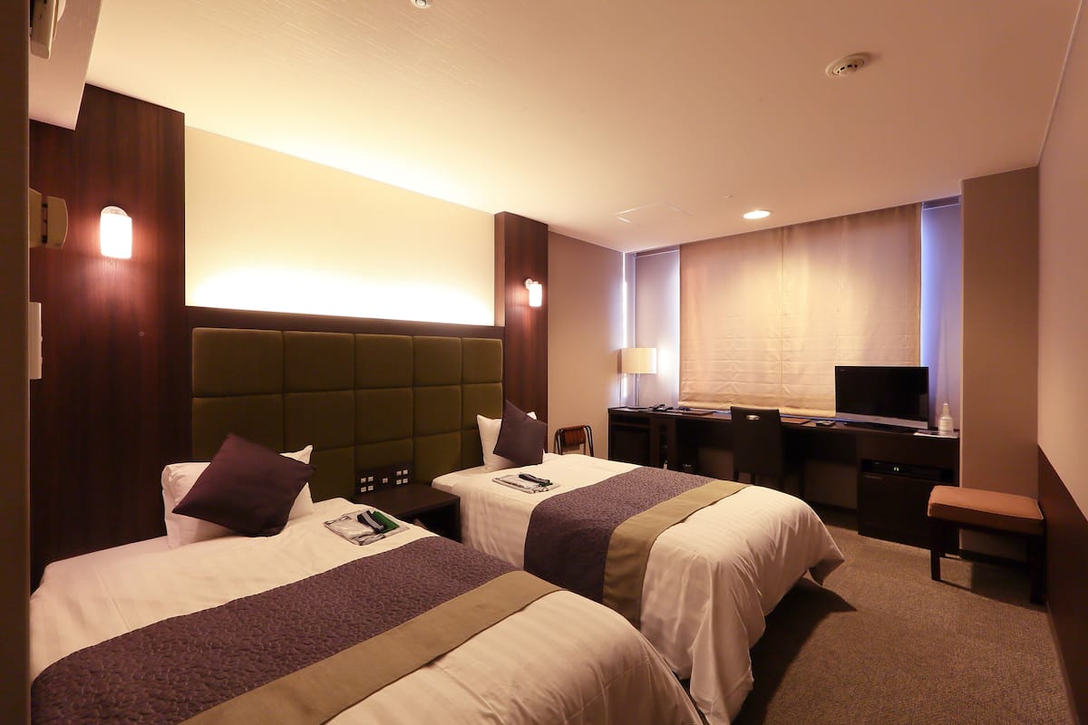 A hotel room with two beds, a desk, a chair, a television, and a lamp.