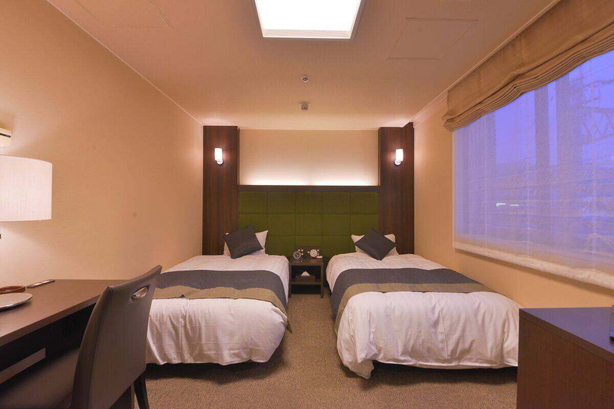 A hotel room with two beds, a desk, and a chair. There is a window with a view of buildings.