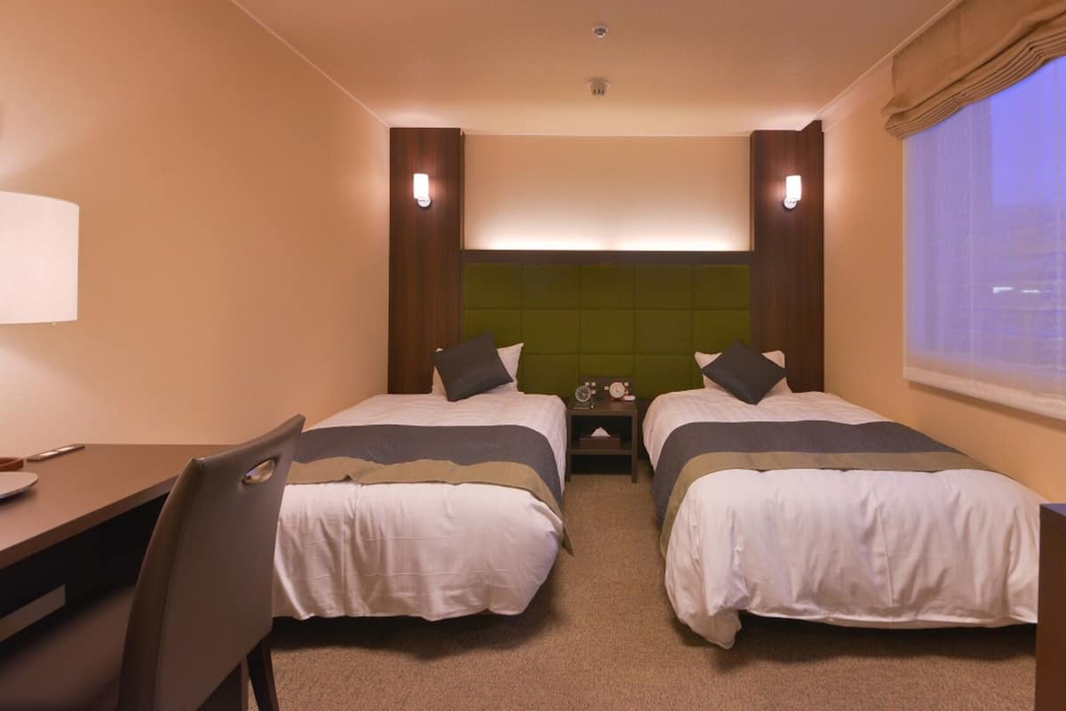 A hotel room with two beds, a desk, a chair, a nightstand, and a window with a view.