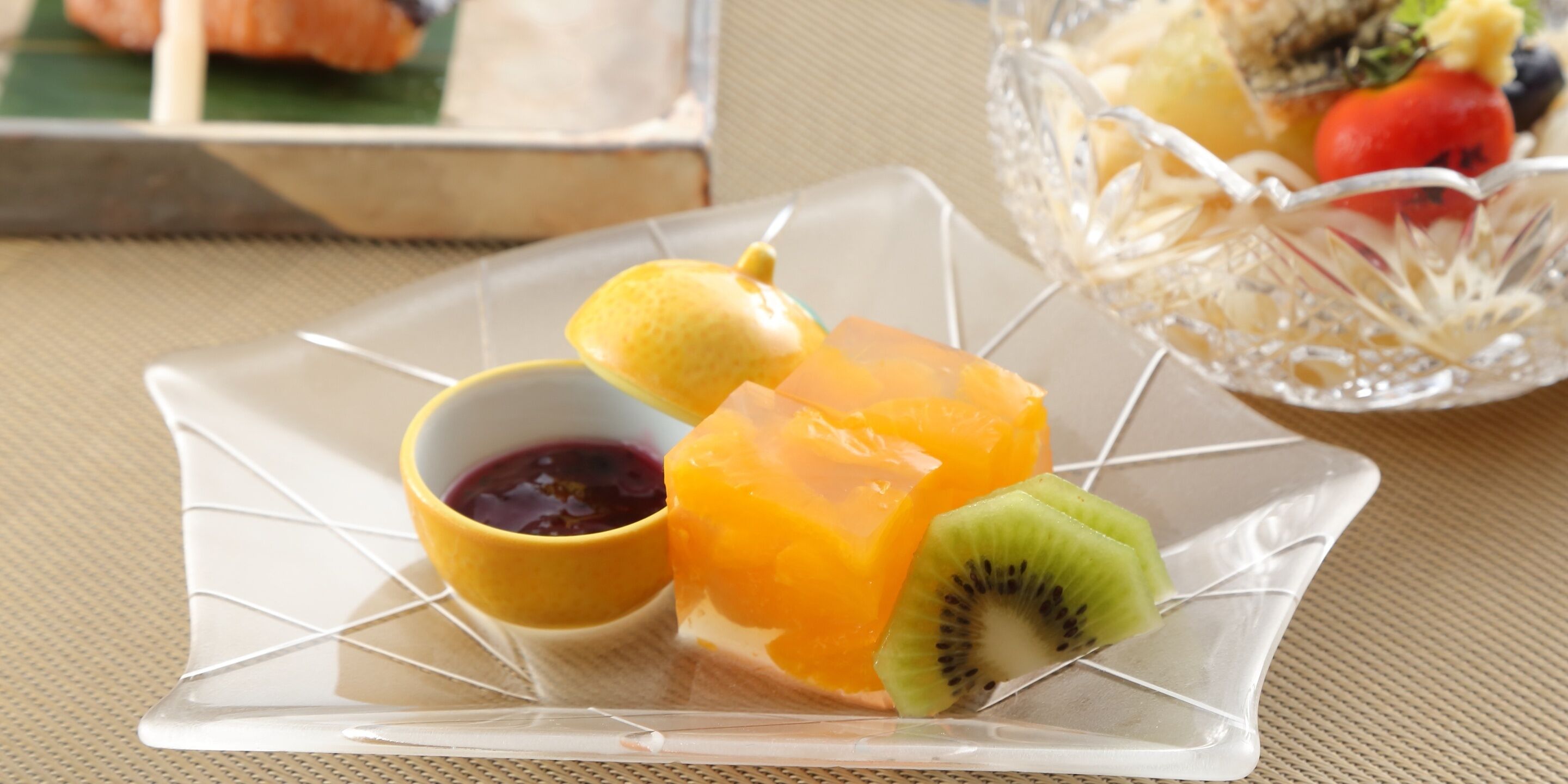Una placa de vidrio transparente con una composición de frutas que incluye kiwi, naranja y un limón, acompañada de un recipiente pequeño con mermelada.