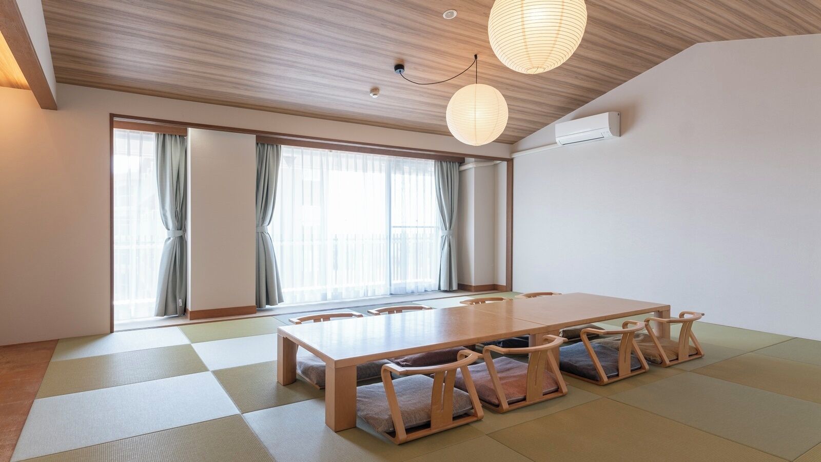 Um quarto com piso de esteiras de tatame, uma mesa de madeira com almofadas e uma grande janela com cortinas.
