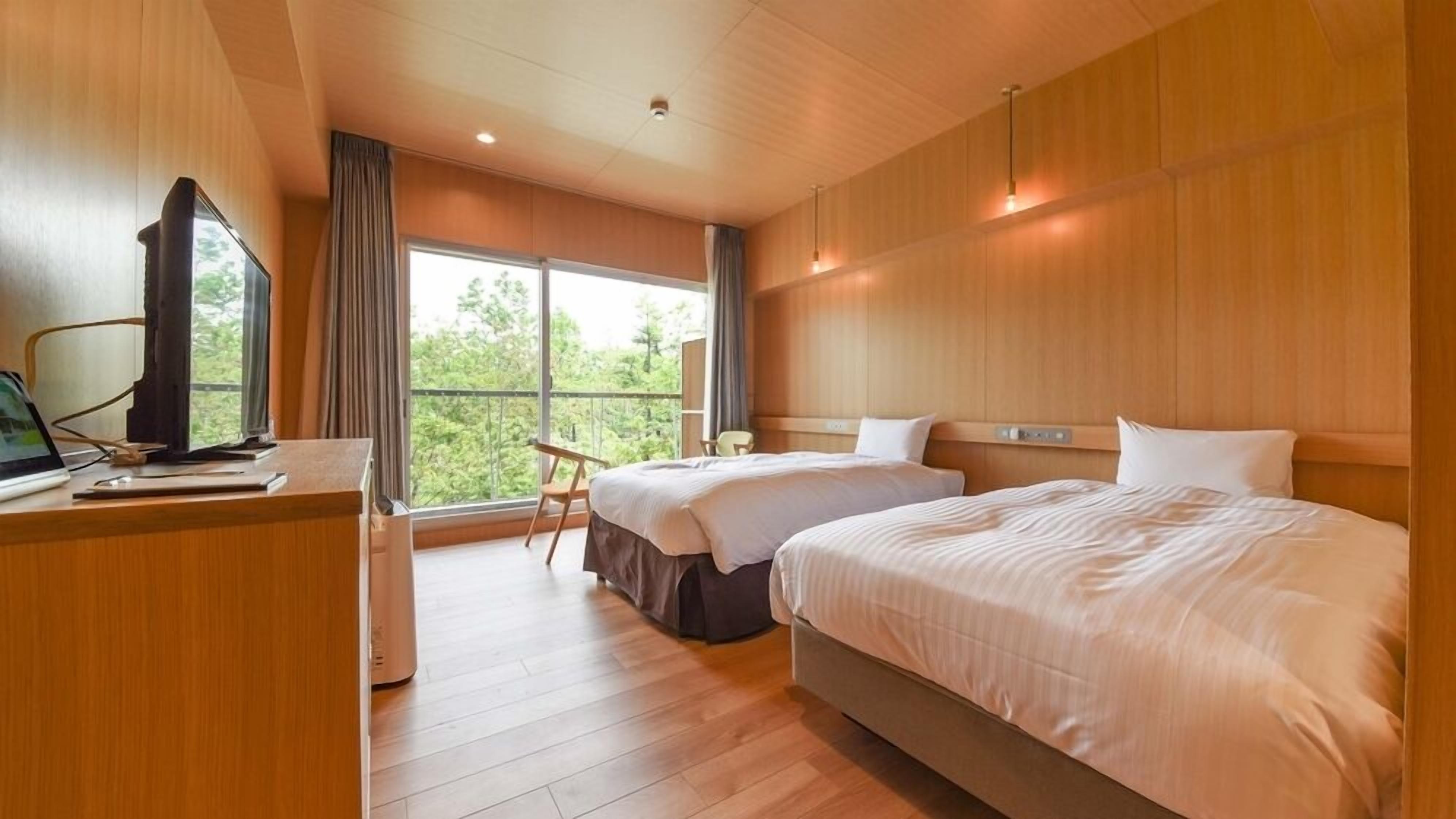 Quarto de hotel com duas camas, uma escrivaninha com computador e uma janela grande com vista para árvores.