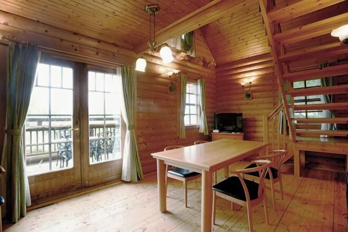 A wooden cabin interior with a dining table, chairs, a TV, and a staircase.