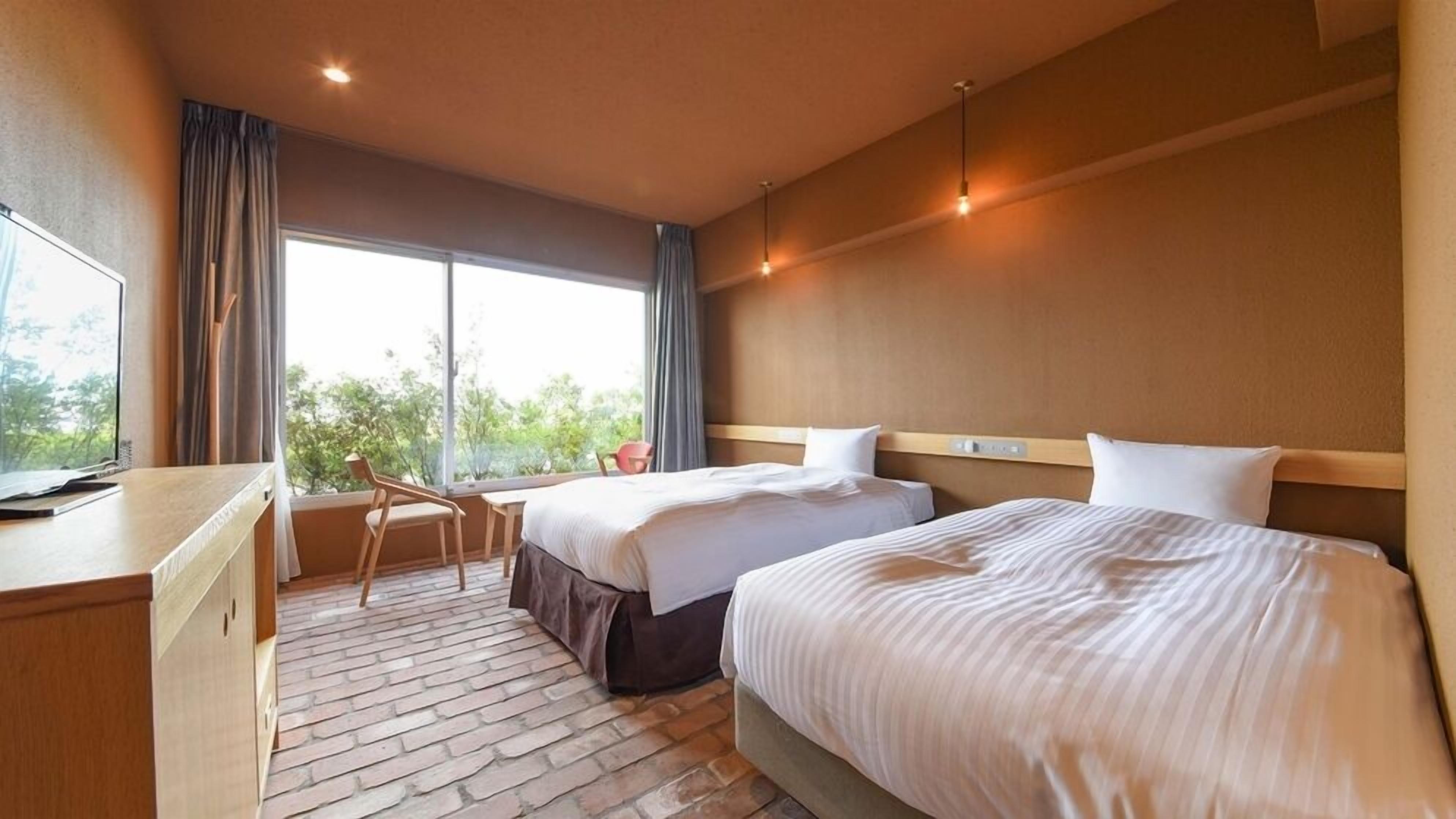 Quarto de hotel com duas camas, uma escrivaninha, uma cadeira, janela com cortinas e vista para o verde lá fora.
