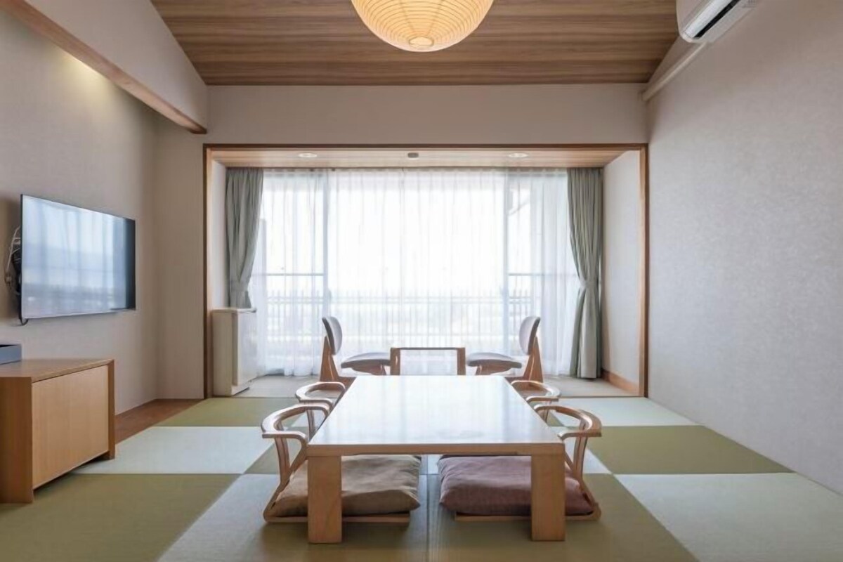 A room with a wooden table, chairs, a TV, and a large window with curtains.