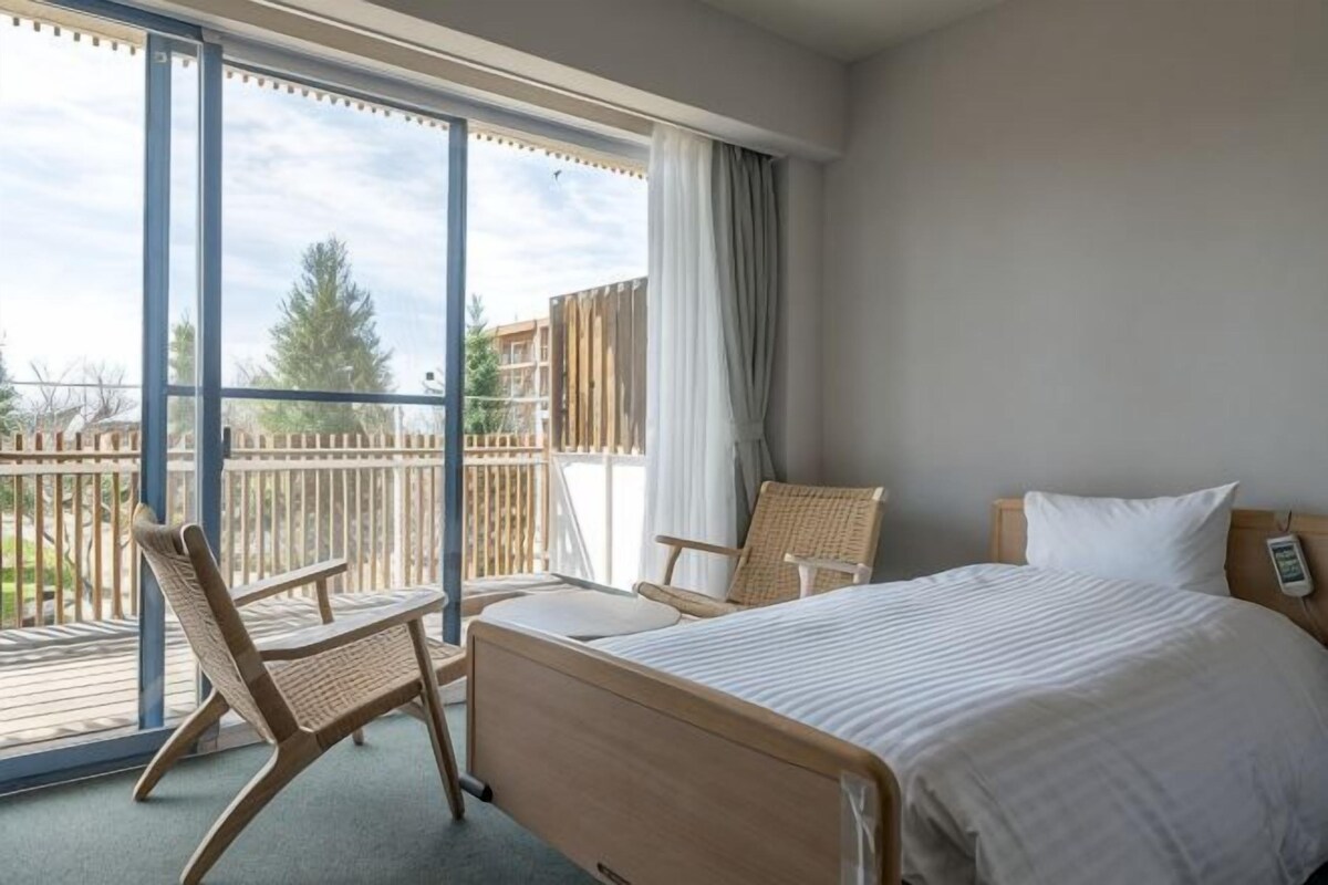 A hotel room with a bed, two chairs, a window, and a balcony.