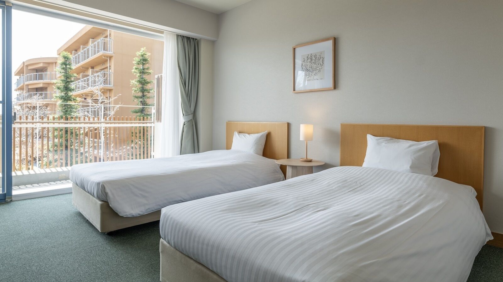 Quarto de hotel com duas camas, janela com vista para um edifício e árvores, uma mesinha com abajur e um quadro na parede.