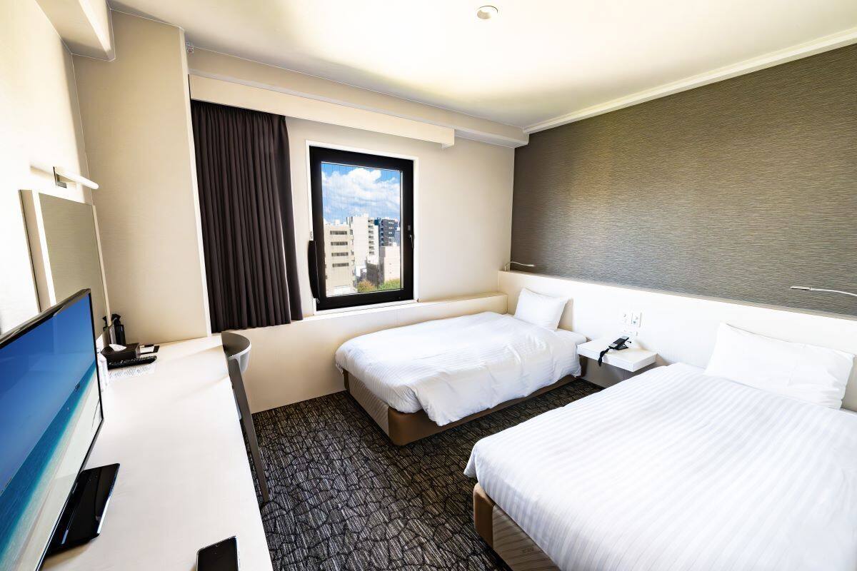 A hotel room with a desk, two beds, a TV, and a window with a city view.