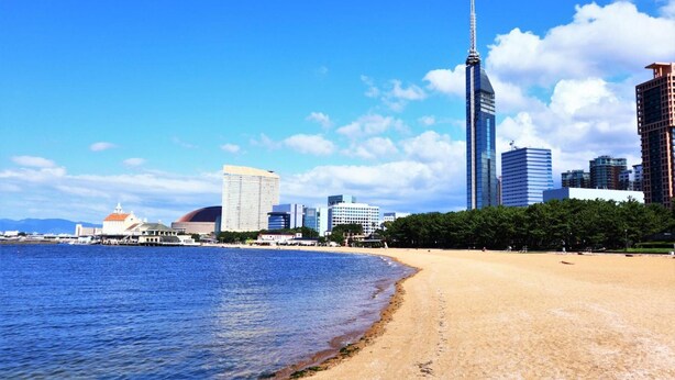Una playa con el horizonte urbano al fondo, incluyendo un rascacielos alto y varios edificios más.