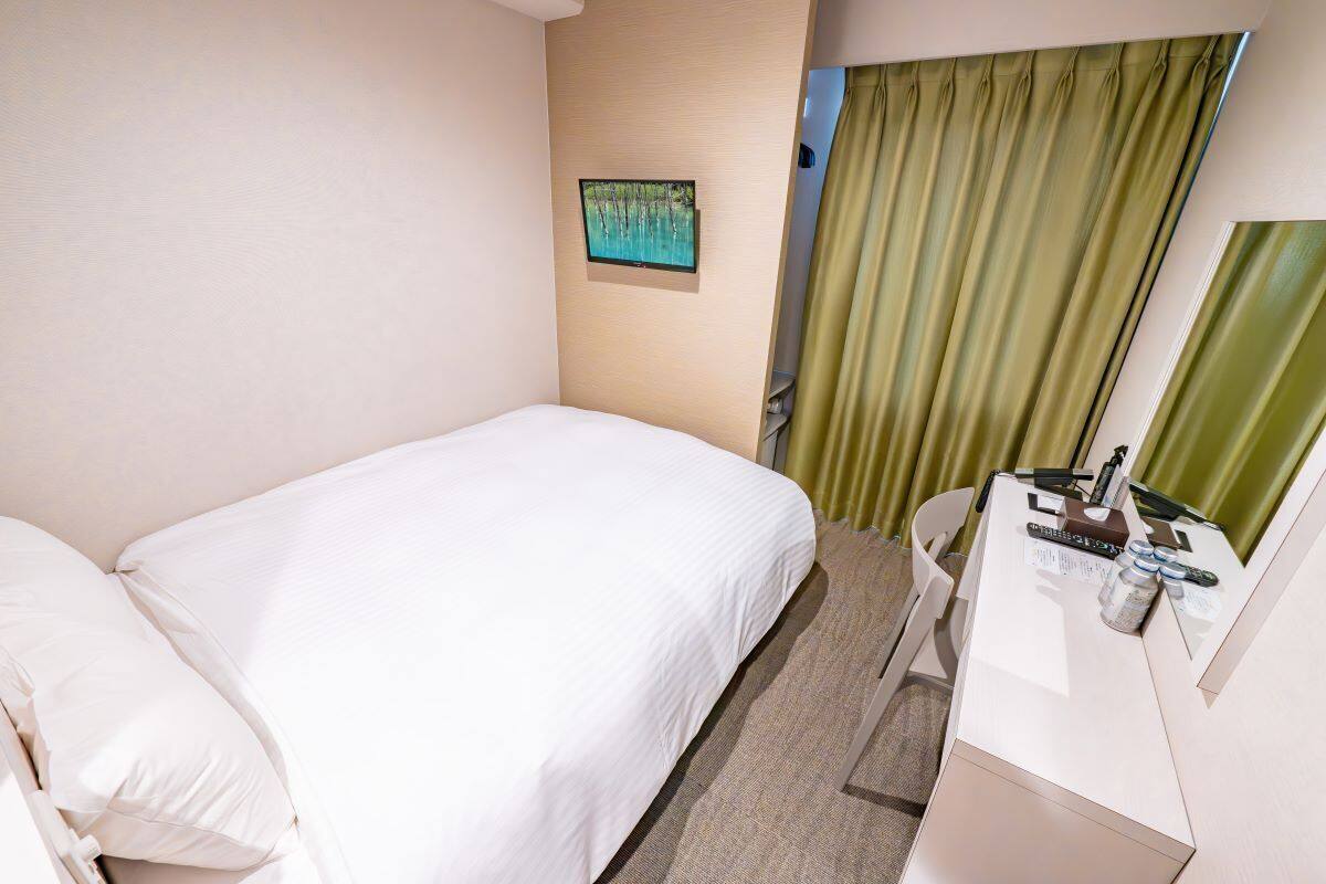 A hotel room with a bed, a desk with a mirror, a television on the wall, and a window with curtains.