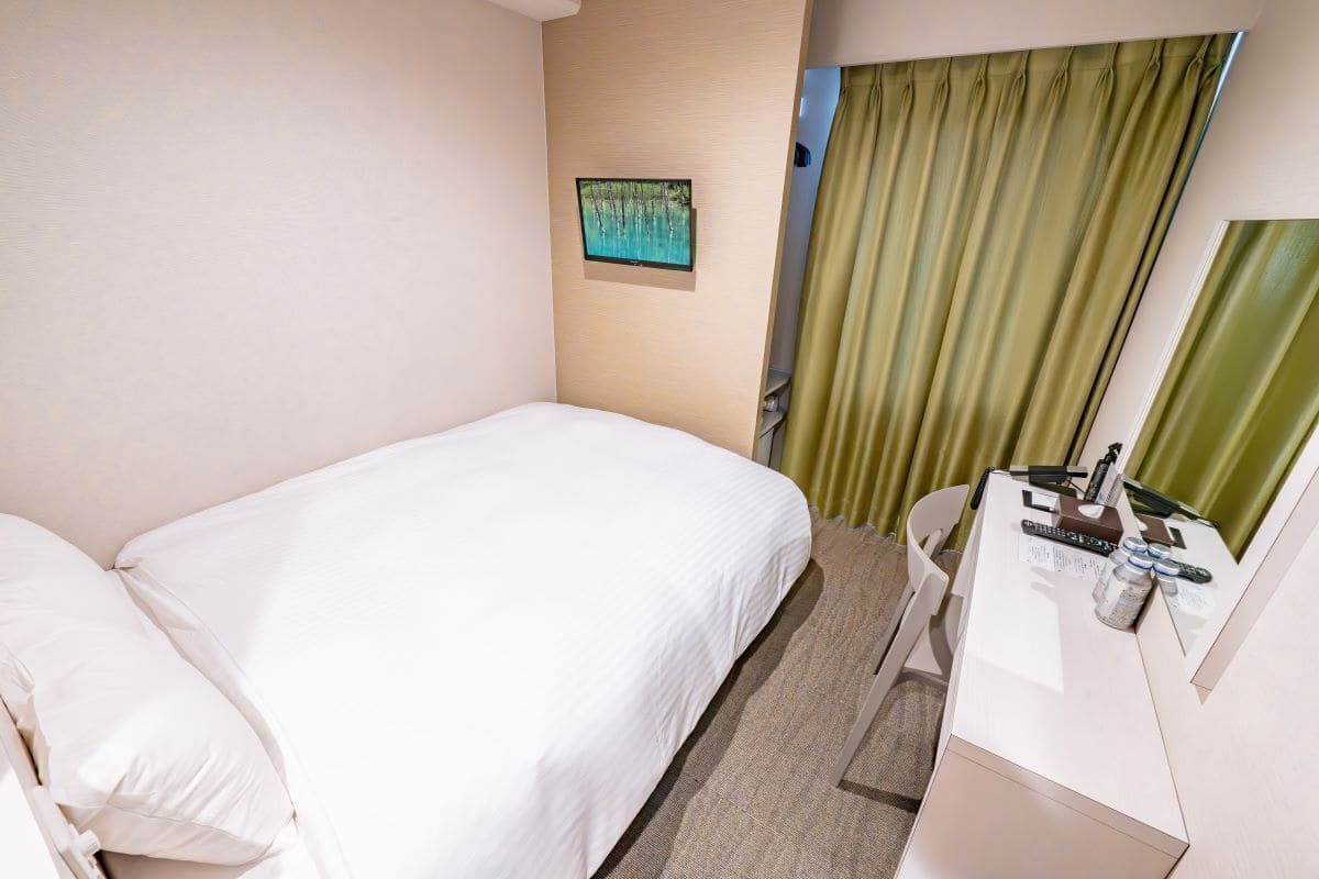 A hotel room with a bed, a desk with a mirror, a television on the wall, and a window with curtains.