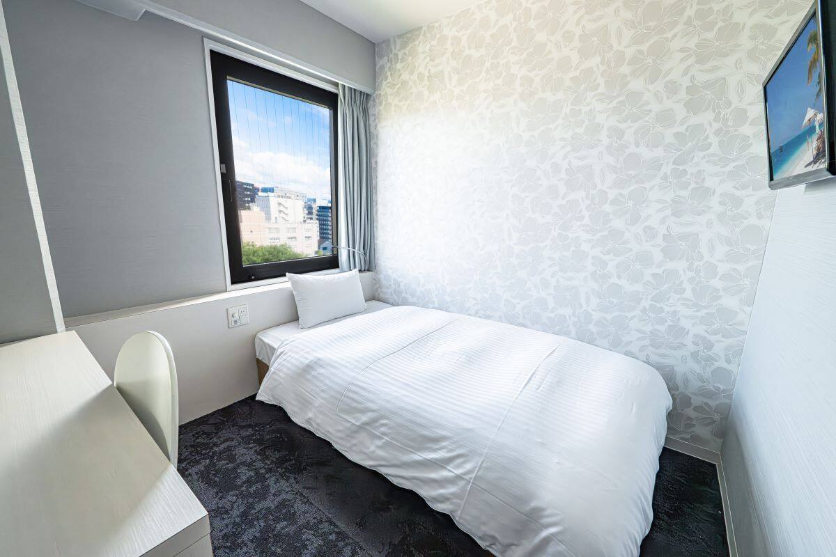 A hotel room with a bed, a desk, a chair, a window with a view of buildings, and a patterned wall.