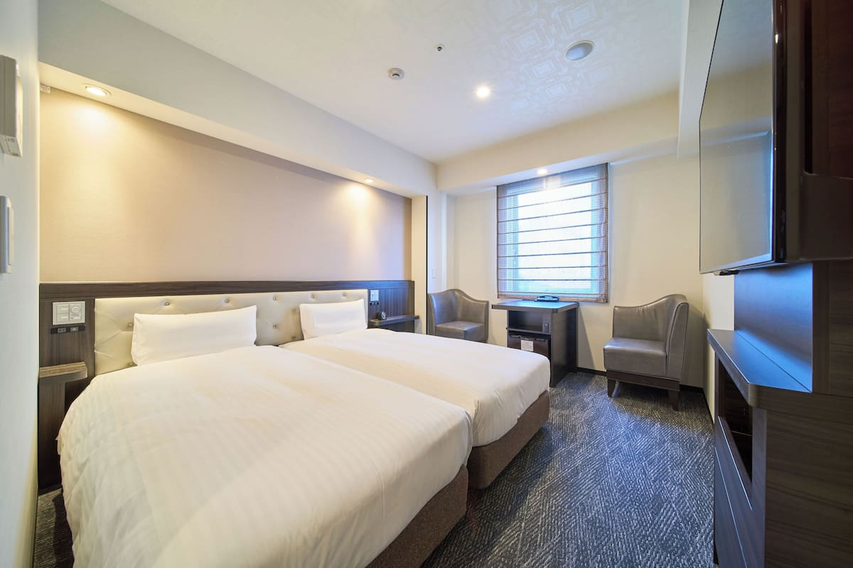 A hotel room with two beds, a desk, a chair, a TV, and a window with blinds.