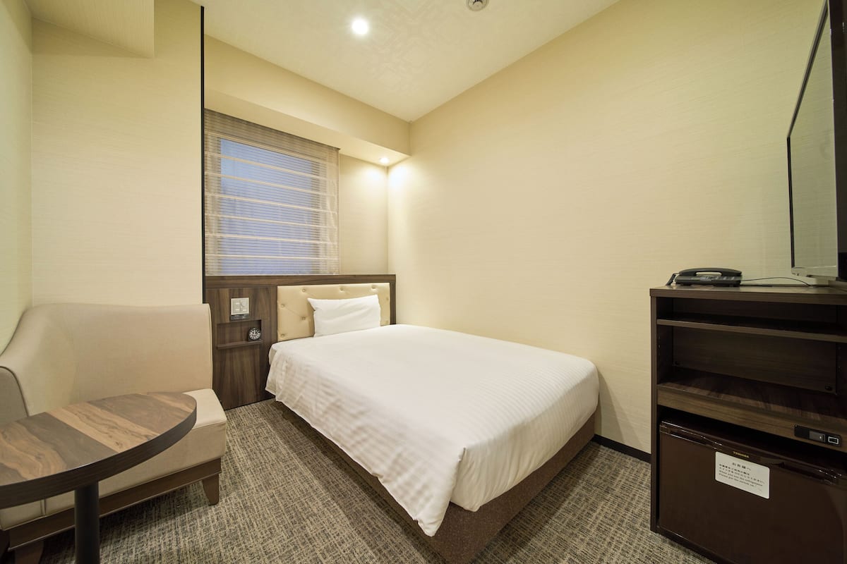 A hotel room with a bed, a small table, a chair, a window with blinds, and a built-in TV.