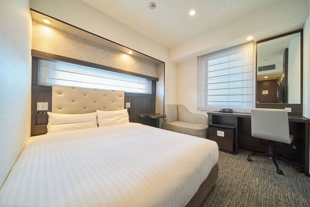 A hotel room with a large bed, a desk with a chair, a mirror, and a window with blinds.