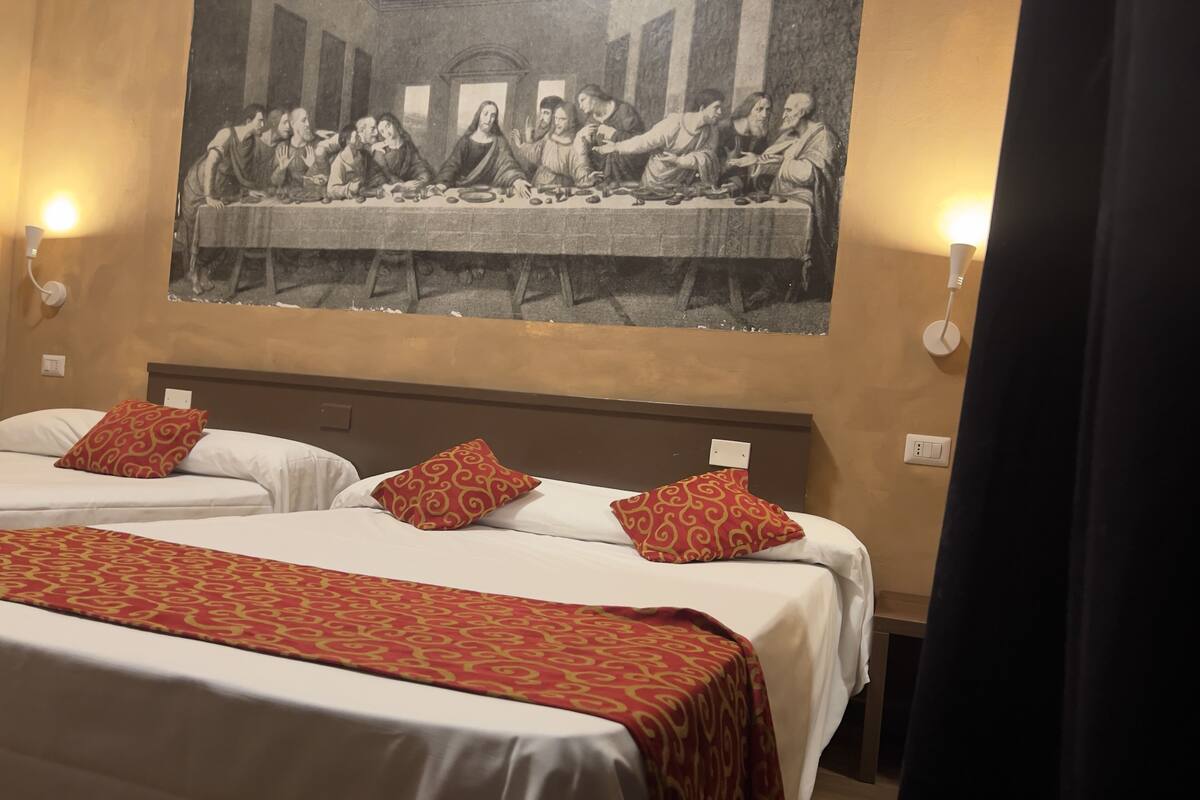 A hotel room with two beds, each with a red patterned bedspread and matching pillows. A large painting of 'The Last Supper' is on the wall above the beds.