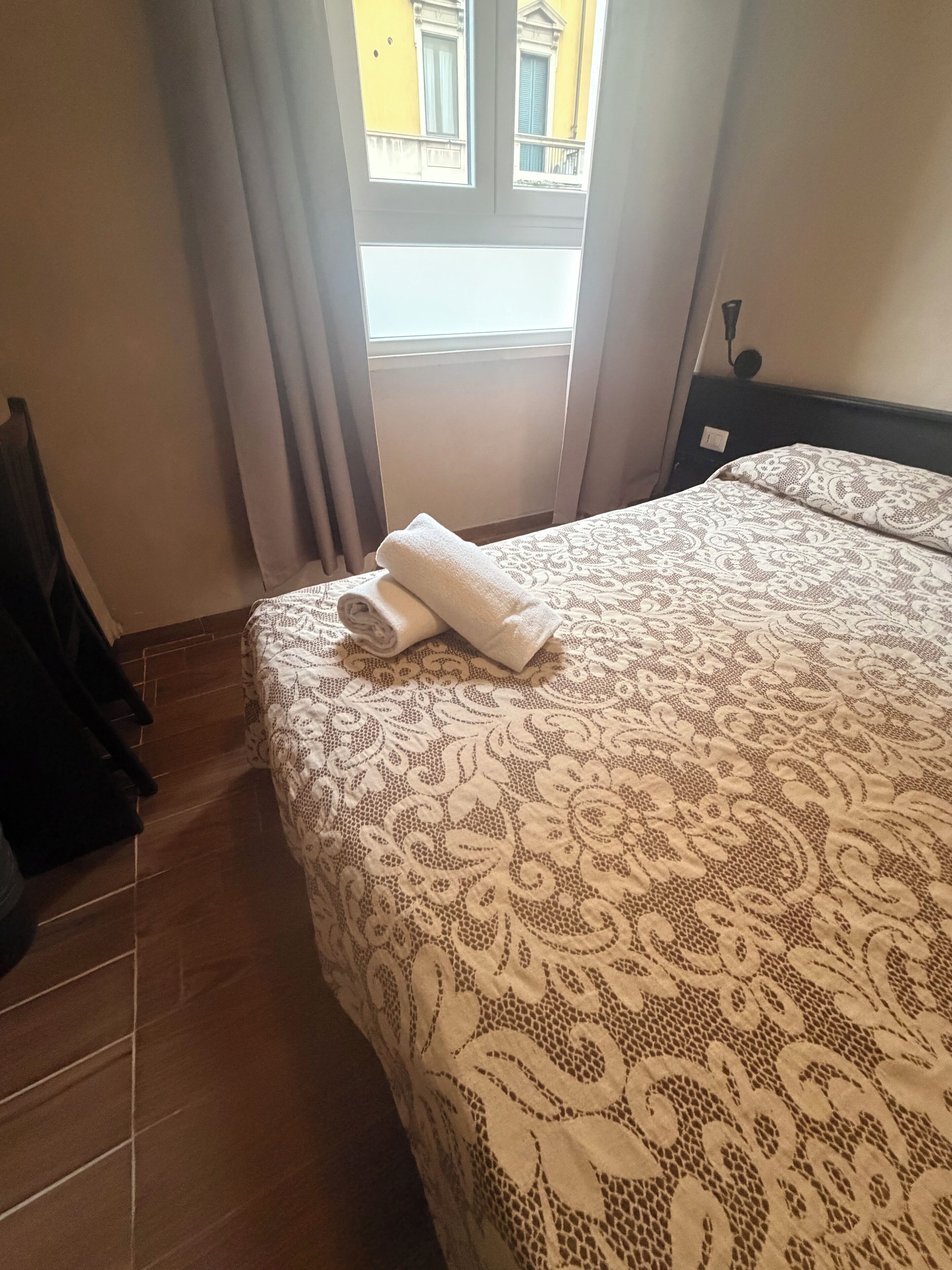 Un lit bien fait, recouvert d’une couvre-lit à motifs, une fenêtre avec des rideaux et un sol carrelé.