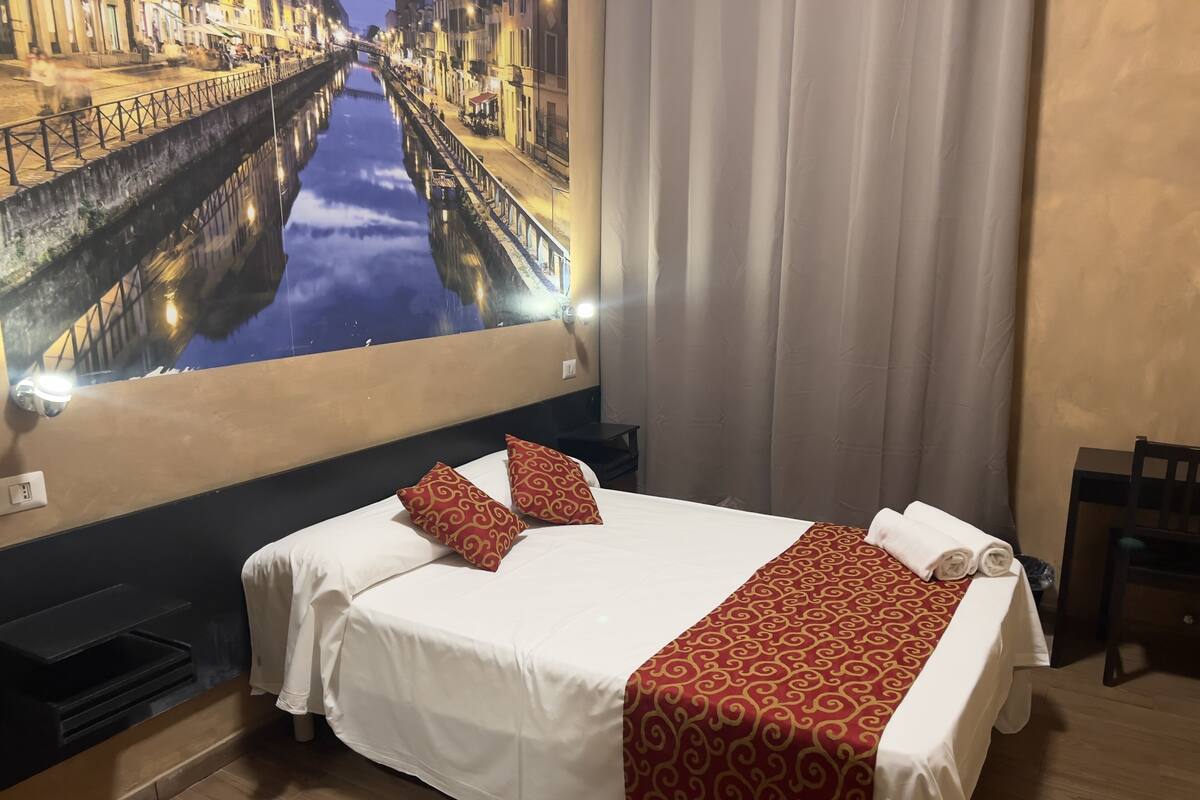 A hotel room with a bed, a nightstand, a chair, and a large window with a cityscape view.