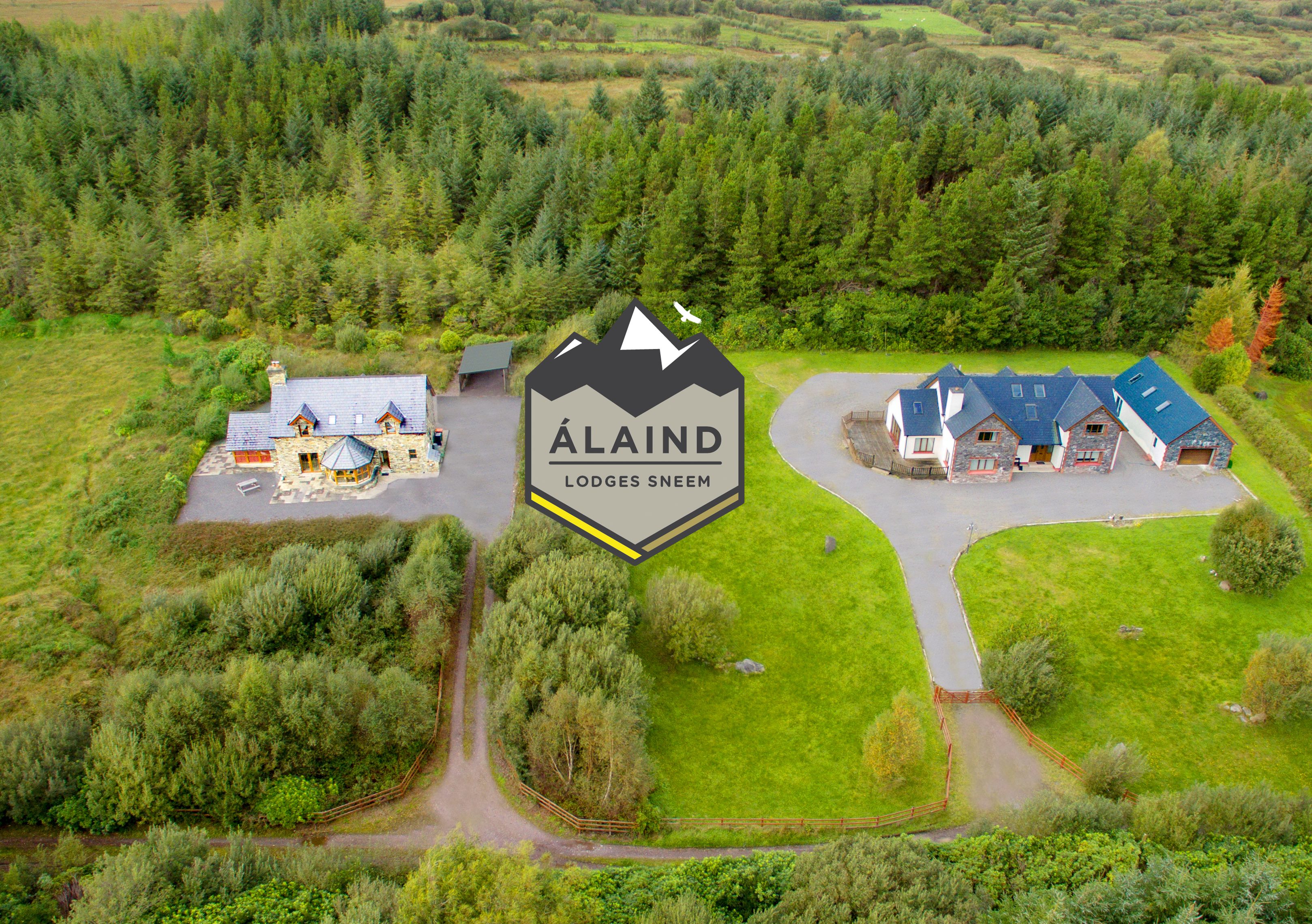 An aerial view of Álaind Lodges Sneeum, featuring a central building with a mountain peak logo, surrounded by lush greenery and multiple guest houses.