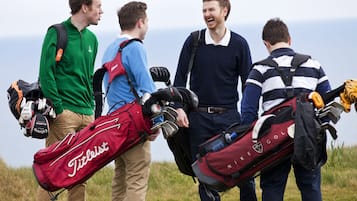 Four people carrying golf bags, standing on a grassy field with a view of the sea in the background.