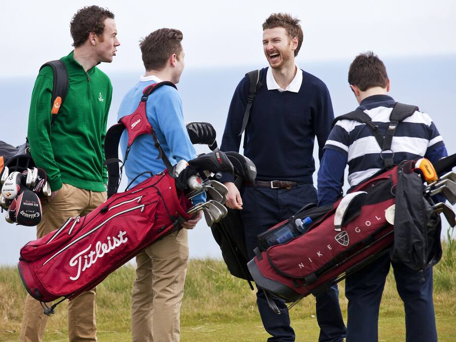 Four people carrying golf bags, standing on a grassy field with a view of the sea in the background.