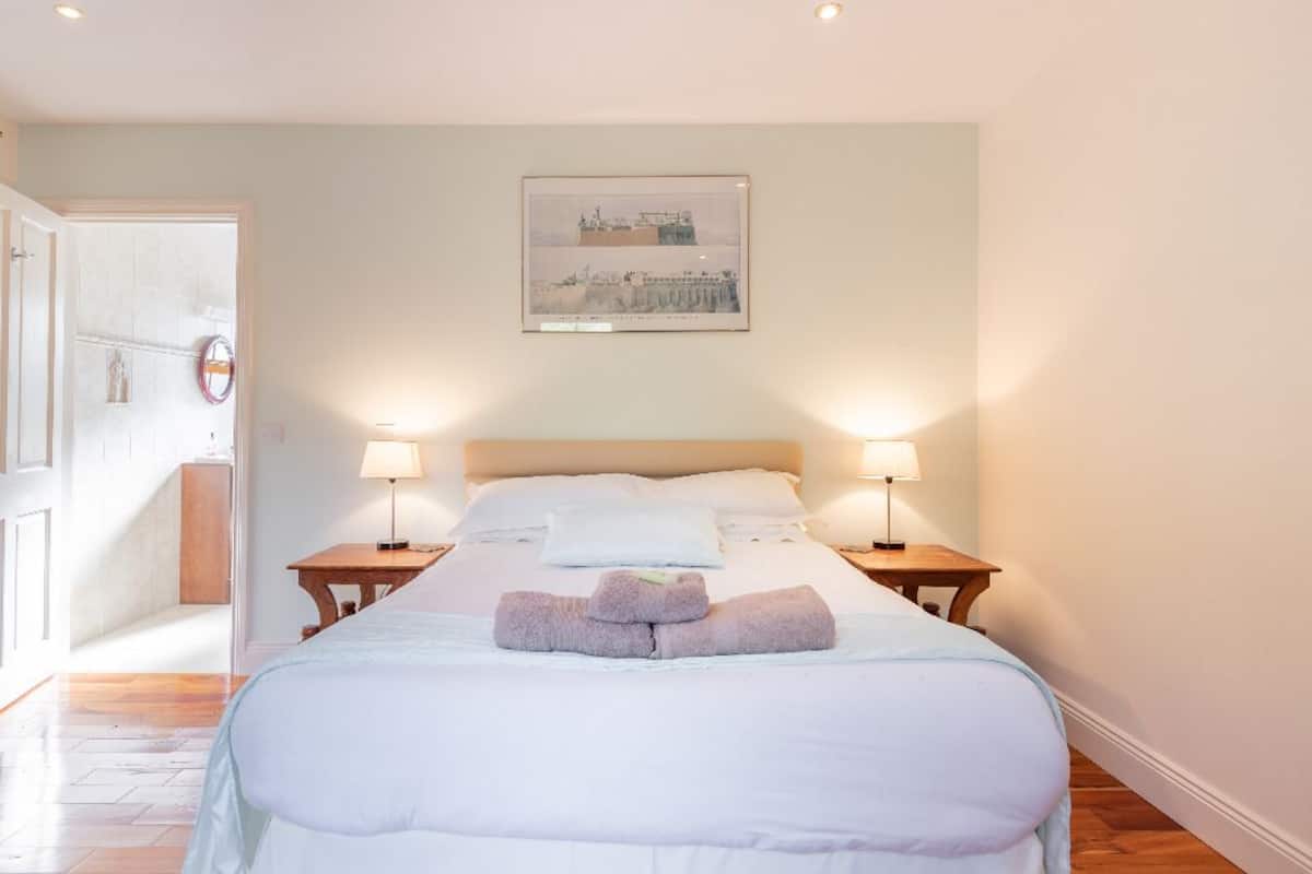 Una habitación de hotel con una cama, dos mesitas de noche con lámparas, un cuadro en la pared y una puerta que da a otra habitación.