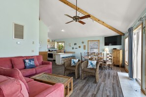Un vaste espace de vie comprenant un canapé, des fauteuils en osier, une table basse, un ventilateur de plafond et, au fond, une cuisine.