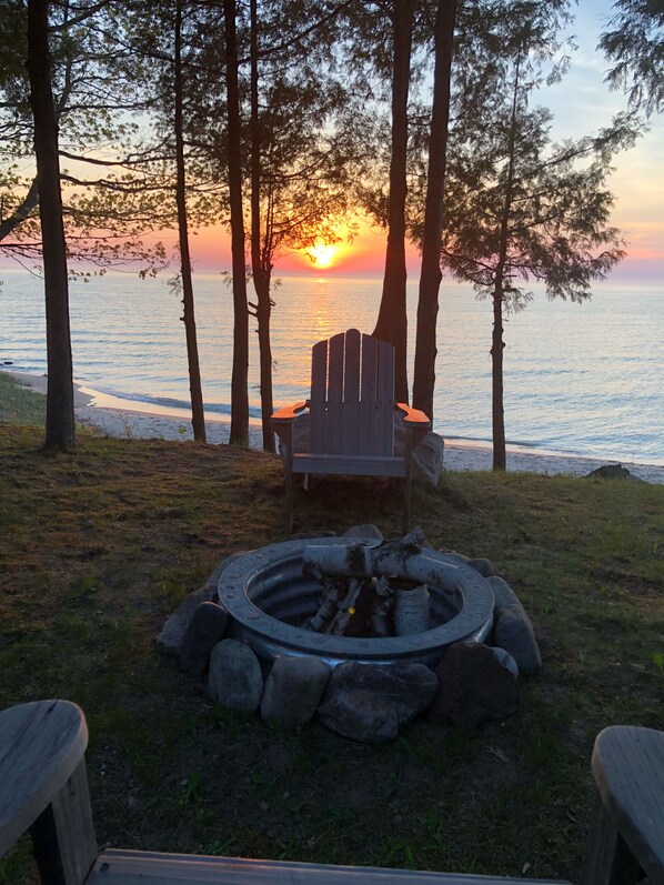 A wooden chair and a fire pit in a natural setting with trees and a view of the sunset over the water.