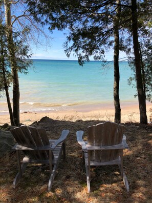 Deux fauteuils Adirondack en bois, placés face à une plage où l’eau est claire et où l’on aperçoit des arbres au loin.