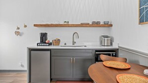 Cocina compacta con lavavajillas integrado, cafetera y tostadora. Encima del lavabo, hay un estante de madera con vasos y una pequeña planta.