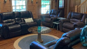 A cozy cabin interior with wooden walls, leather sofas, a glass coffee table, and a wooden staircase. - Indulge in the ultimate vacation experience at this exquisite custom log home.  (Oak Bluffs)