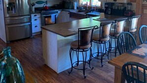 A spacious kitchen with wooden cabinets, a central island with bar stools, stainless steel appliances, and a large window providing natural light. - Indulge in the ultimate vacation experience at this exquisite custom log home.  (Oak Bluffs)